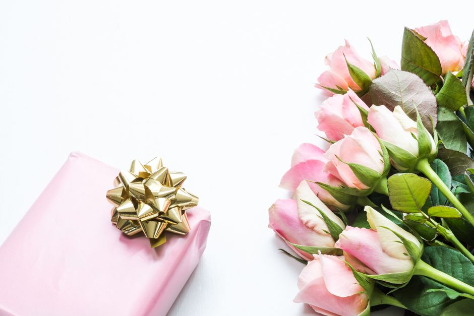 A neatly wrapped pink gift box adorned with a shiny gold bow is positioned on a white surface next to a bouquet of fresh, pale pink roses with green leaves. The roses are partially open with soft peta