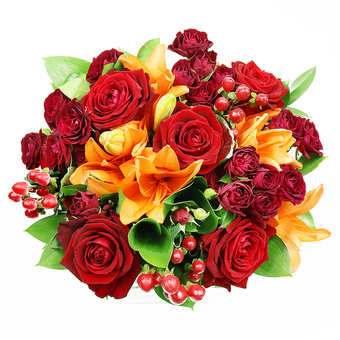 From a bird's-eye perspective against a spotless white backdrop, this luxurious round bouquet reads as an opulent tableau of reds, oranges and fresh greens, meticulously layered to create depth and movement. Large, fully bloomed deep red roses form the heart of the design, their velveteen petals rolling outward in sensuous curves that speak of classic romance and tactile richness. Bright orange Asiatic lilies punctuate the arrangement with trumpet-shaped silhouettes and visible stamens; some stand wide open and bold, their inner markings adding delicate contrast, while others remain elegantly furled, suggesting a bouquet that will continue to evolve after gifting. Tucked between the larger focal flowers are dense clusters of deep burgundy spray roses, their miniature blooms contributing a compact, sumptuous texture and intensifying the jewel-toned dimension. Scattered throughout, glossy hypericum berries-small and spherical, the colour of ripe cranberries-catch the light and offer lively, reflective points against the matte petals. Generous green foliage, likely ruscus or salal, frames and lifts the composition with broad leaves that refresh the warm tones and create a natural, garden-fresh silhouette. The soft, even lighting reveals subtle surface variations-the lilies' satin glow versus the roses' plush nap-while evoking a faint scent of spicy citrus and classic rose perfume. In mood the bouquet suggests celebration and deep affection, well suited to anniversaries, romantic gestures, or seasonal autumn décor. It's the kind of arrangement neighbours in Bow and visitors from nearby Mile End would recognise as a considered, heartfelt choice for marking special moments.