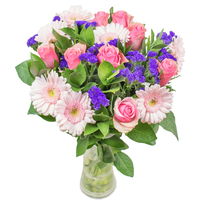 A visually stunning floral arrangement, presented in a crystal-clear glass vase on a crisp white background, captures the welcoming spirit of Bow. This standout display features approximately seven to nine soft to medium pink roses, their petals elegantly curled in full or partial bloom, echoing the classic rose spiral. Intertwined are five to seven blush gerbera daisies, each with a broad, gentle face and delicate, radiating petals, their pale pink tones almost ethereal next to creamy-white centers. Interspersed throughout are lush clusters of purple statice, adding lively pops of colour and intriguing texture-much like the diversity found in Bow's vibrant market stalls. Deep green, plentiful foliage encircles the blooms, providing structure, freshness, and echoes of nature along the River Lea towpath. The underlying glass vessel reveals tidy stems and clear water, reinforcing a sense of natural beauty-perfect for local gifting or delivery across Bow. This versatile assortment, suitable for occasions like birthdays, anniversaries, or Mother's Day, offers a joyful, affectionate tribute, radiating the careful artistry and warmth of an East London florist.